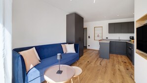 A modern living space with a blue sofa, a small round table, and a kitchen area with grey cabinets and a wooden countertop.