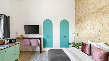 A bedroom with a bed, a desk with a computer, two chairs, a TV, and a stone wall.