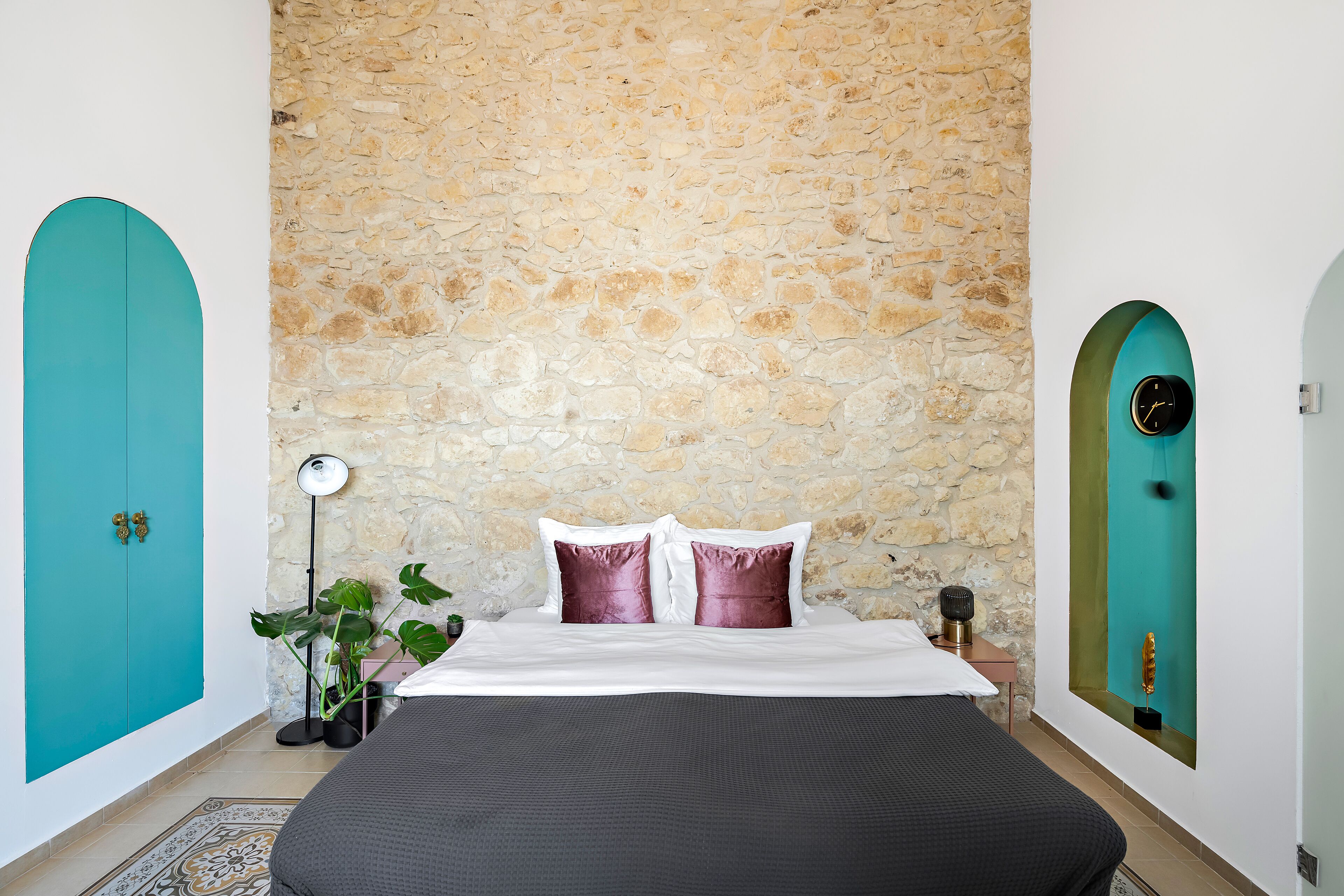 A bedroom with a stone wall, teal doors, a bed with white and purple pillows, a floor lamp, and a potted plant.