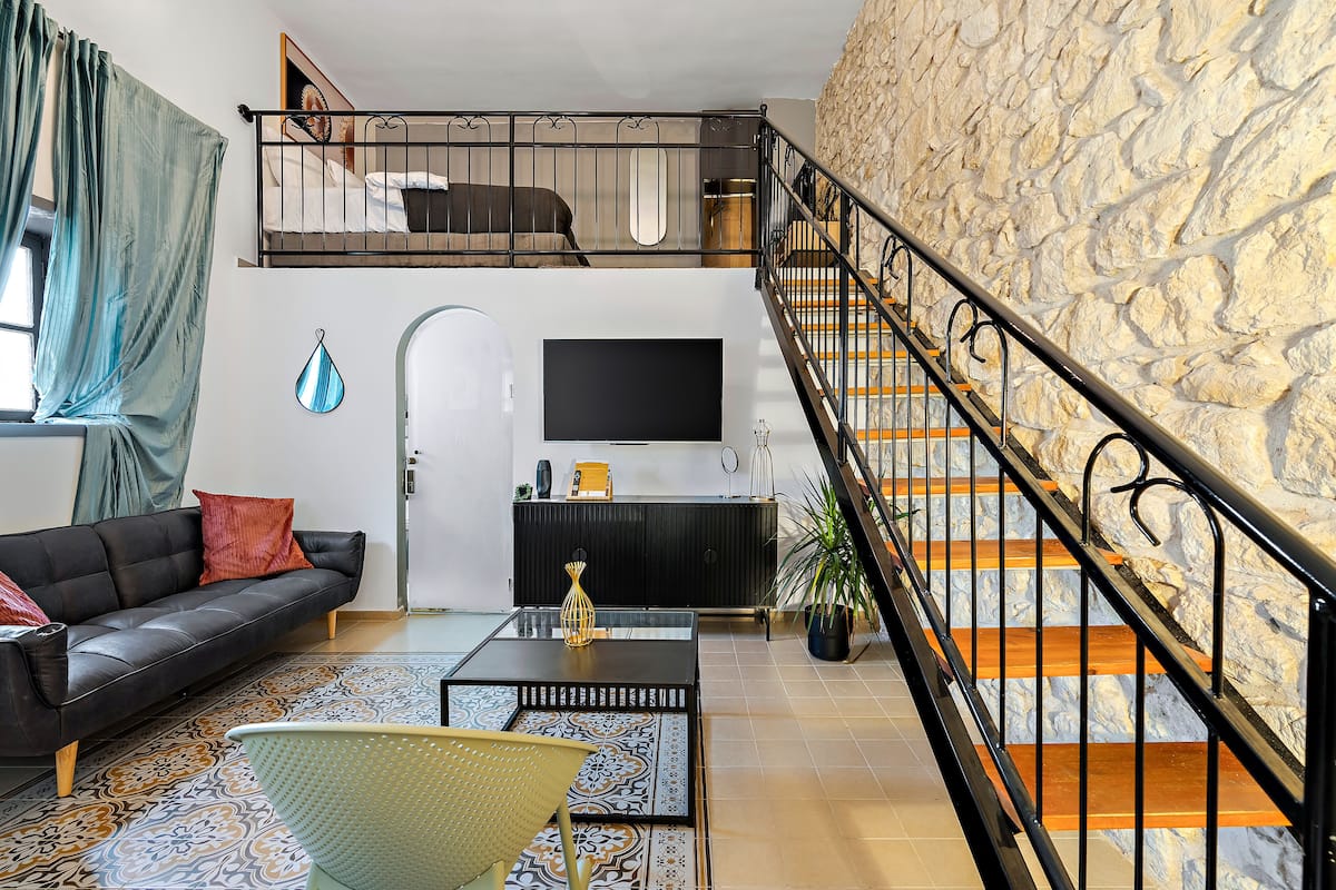 Ein modernes Wohnzimmer mit einer Couch, einem Couchtisch und einem Fernseher. Eine Treppe mit einer Steinwand und ein Balkon darüber.