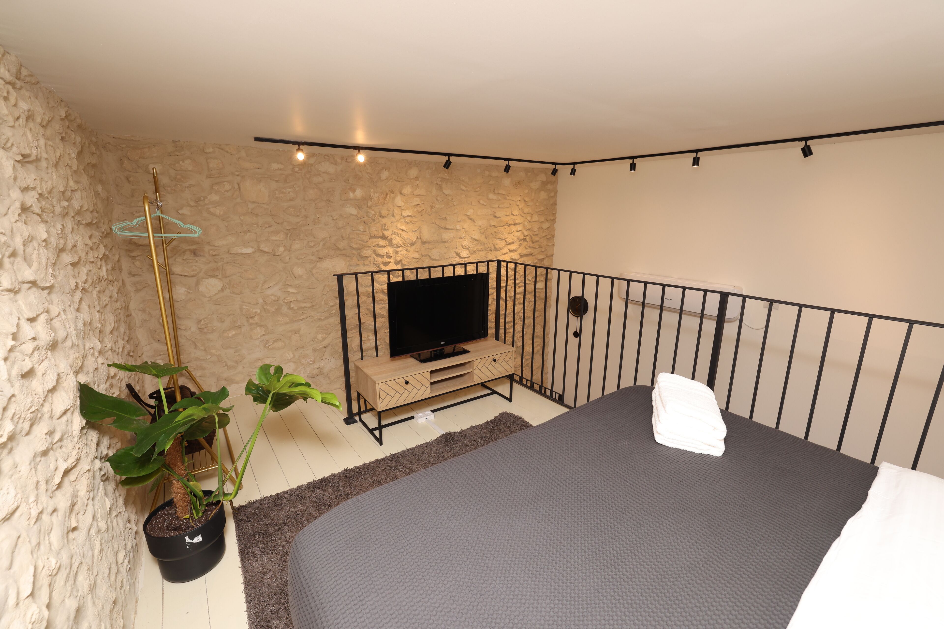 A bedroom with a bed, a TV on a wooden stand, a potted plant, and a stone wall.