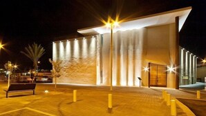 A modern building with a large entrance, illuminated by exterior lights, and a paved area with a bench and bollards. - Helios Boutique Suites by La Finca Hotel (Beersheba)