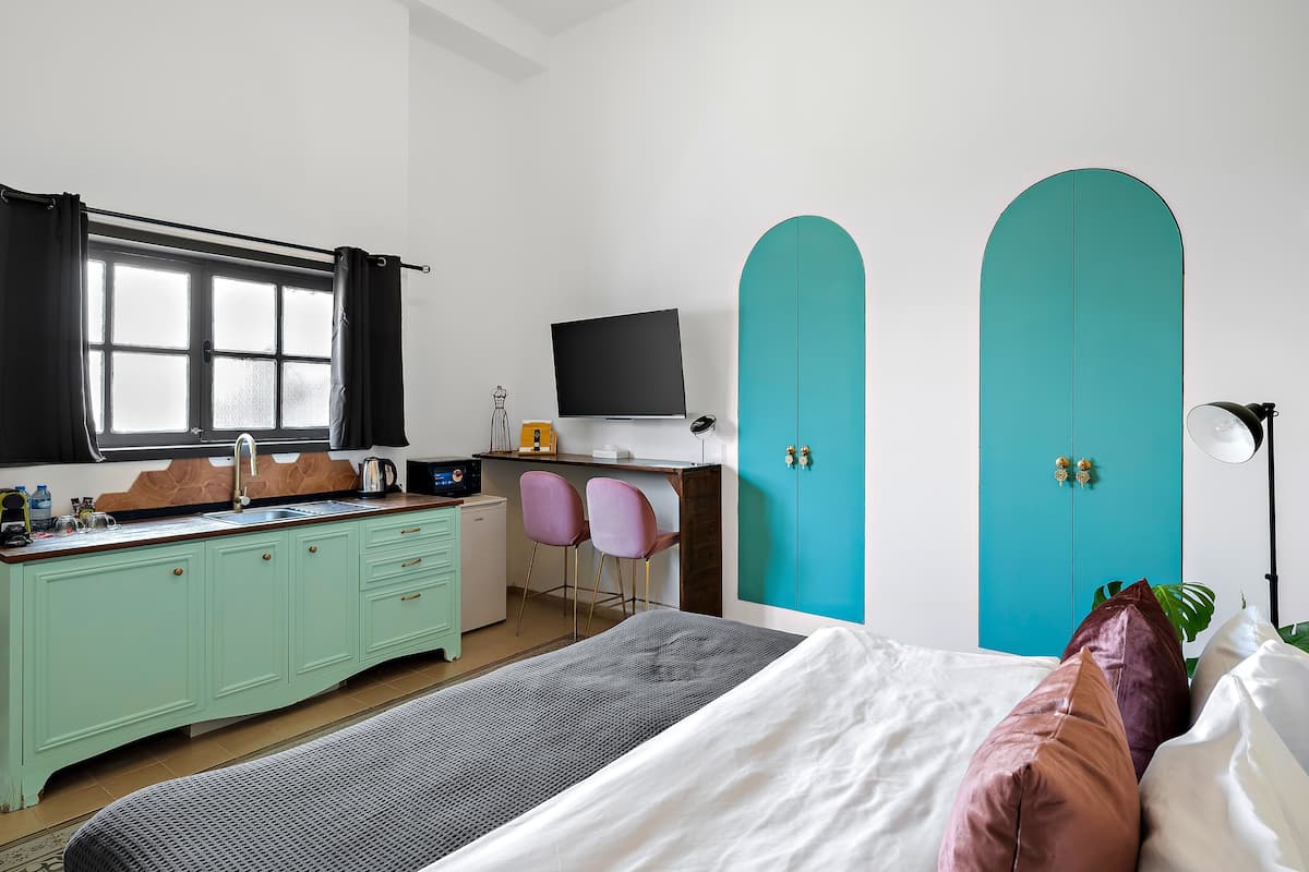 Ein Zimmer mit Bett, Schreibtisch, Fernseher, einem Fenster mit Vorhängen, einer Küchenzeile und türkisblauen Schrankschränken.