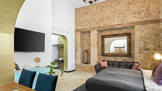 A modern bedroom with a stone wall, a large bed, a flat-screen TV, and a dining area with a wooden table and chairs. - Helios Boutique Suites by La Finca Hotel (Beersheba)