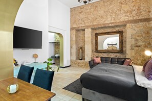 A modern bedroom with a stone wall, a large bed, a flat-screen TV, and a dining area with a wooden table and chairs. - Helios Boutique Suites by La Finca Hotel (Beersheba)