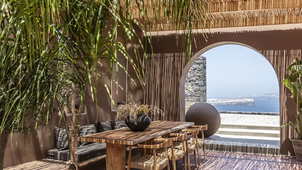 An outdoor seating area with a wooden table and chairs, a stone pillar, and a view of the sea. - Serra Exclusive Suites (Santorini)