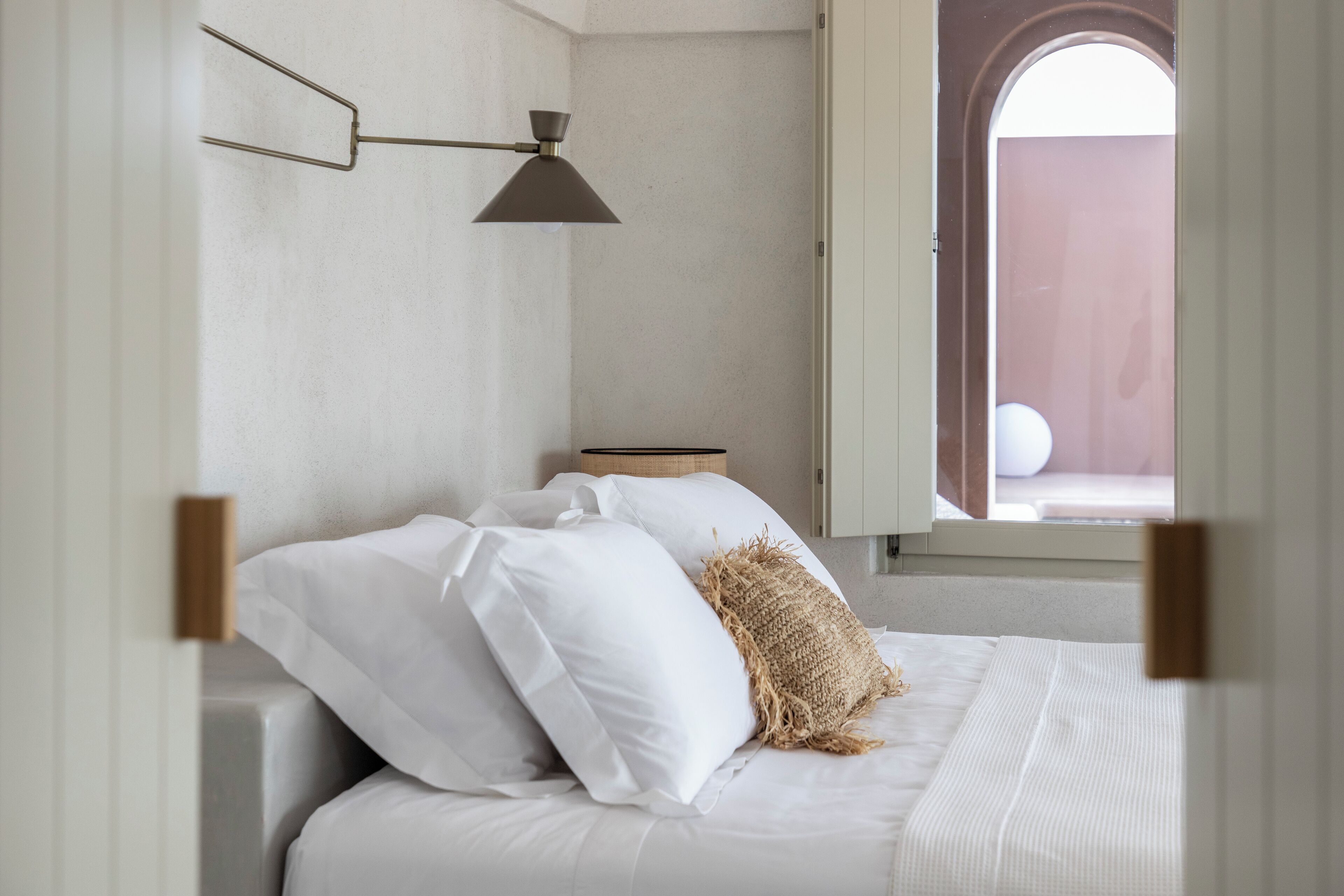 A bedroom with a bed, white bedding, a hanging lamp, and a view through an open window.