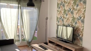 Una sala de estar moderna con un sofá, una mesa de centro, un televisor sobre un soporte de madera y un ventanal con cortinas.