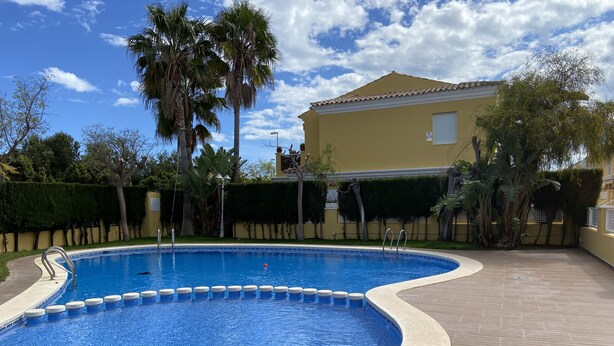 Una piscina con borde de azulejos, rodeada por una terraza de madera, palmeras y un edificio con techo de tejas.