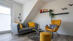 Ein modernes Wohnzimmer mit grauem Sofa, gelbem Sessel und Pouf, einem Glastisch und einem kleinen Teppich.