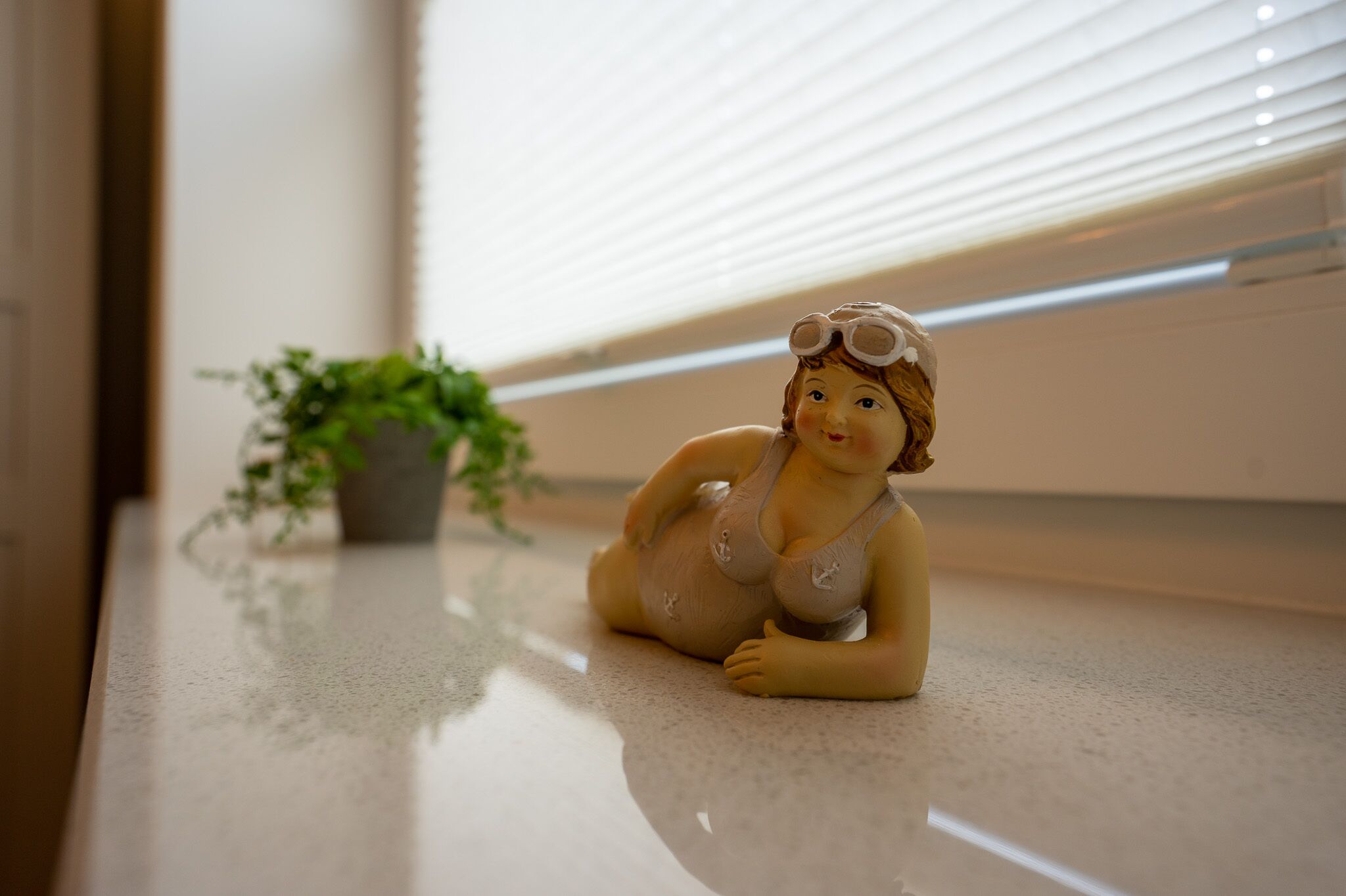 A figurine of a child in a swimsuit with goggles on, placed on a polished floor near a window with blinds.