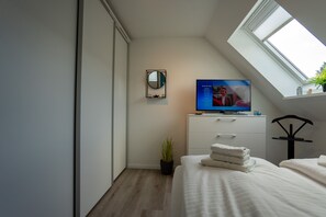 Quarto moderno com claraboia, televisão, cama com lençóis brancos, uma pequena planta em vaso e cabeceira preta.