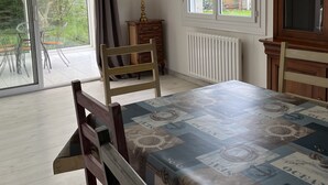 A dining area with a table covered in a patterned tablecloth, a wooden chair, a radiator, and a window with a view of a house and greenery.