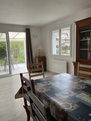 Ein Essbereich mit Tisch, bedeckt mit einer gemusterten Tischdecke, einem Holzstuhl, einem Heizkörper und einem Fenster mit Blick auf ein Haus und Grünflächen.