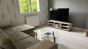 A living room with a beige sofa, a glass coffee table, a TV stand with shelves, and a window with a view of greenery.