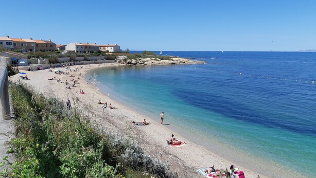 Ein Strand mit klarem, blauem Wasser, Menschen, die sich sonnen, und Gebäuden im Hintergrund.