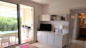 Ein modernes Wohnzimmer mit einer Glasschiebetür, die auf einen Balkon führt, einem Flachbildfernseher, einem kleinen Tisch mit rosa Unterlagen und einer weißen Kommode mit Büchern und Dekorationsobjekten.
