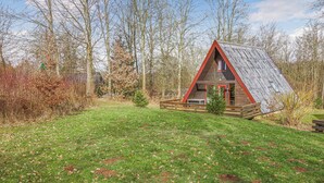 Une petite cabane triangulaire en bois, avec un toit rouge et une terrasse en bois, entourée d’arbres et d’herbe.