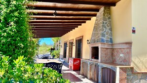 An outdoor patio with a stone fireplace, a Coca-Cola cooler, and a covered seating area with a view of the surrounding greenery. - Finca La Panorámica (Málaga)