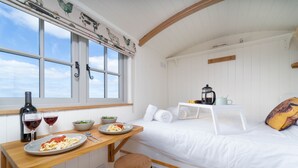 Una habitación acogedora con cama, una mesita con una botella, vasos y comida, además de una ventana con cortinas.