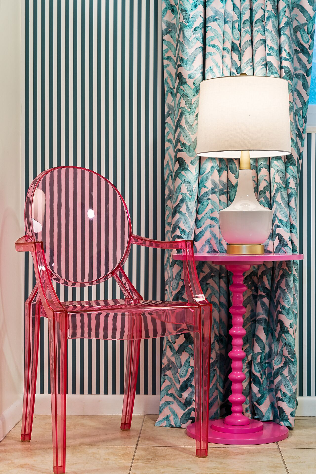 Een kamer met een gestreepte wand, een stoel met een transparant roze frame, en een tafel met een roze voet en een lamp.