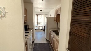 Una cocina estrecha con armarios de madera, un refrigerador y un comedor a la vista al fondo.