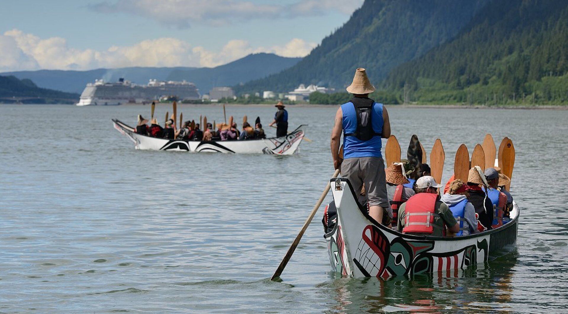 Una canoa tradicional con pasajeros, una persona remando y otra canoa al fondo.
