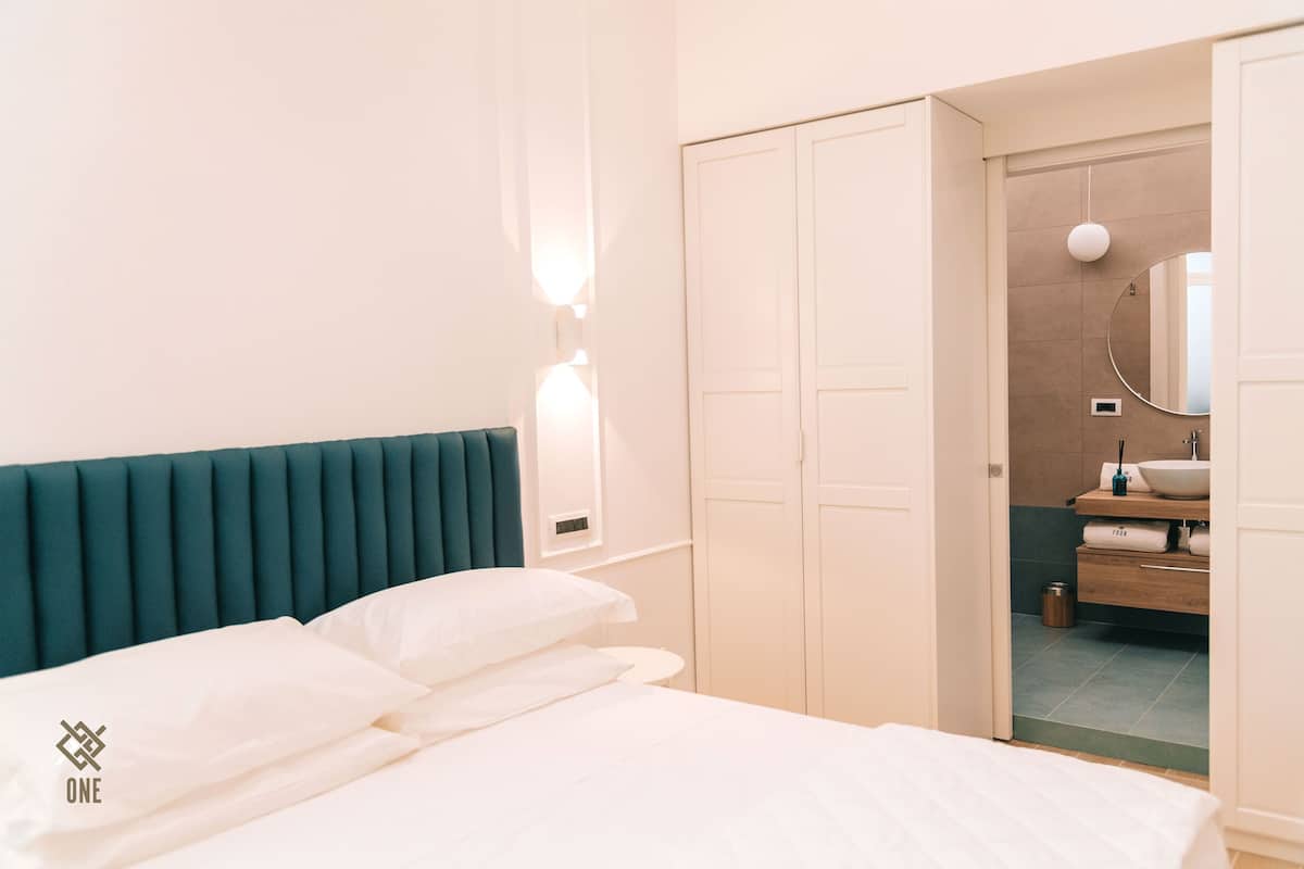 A hotel room with a bed, a teal headboard, a white wall, and a bathroom visible through an open door.