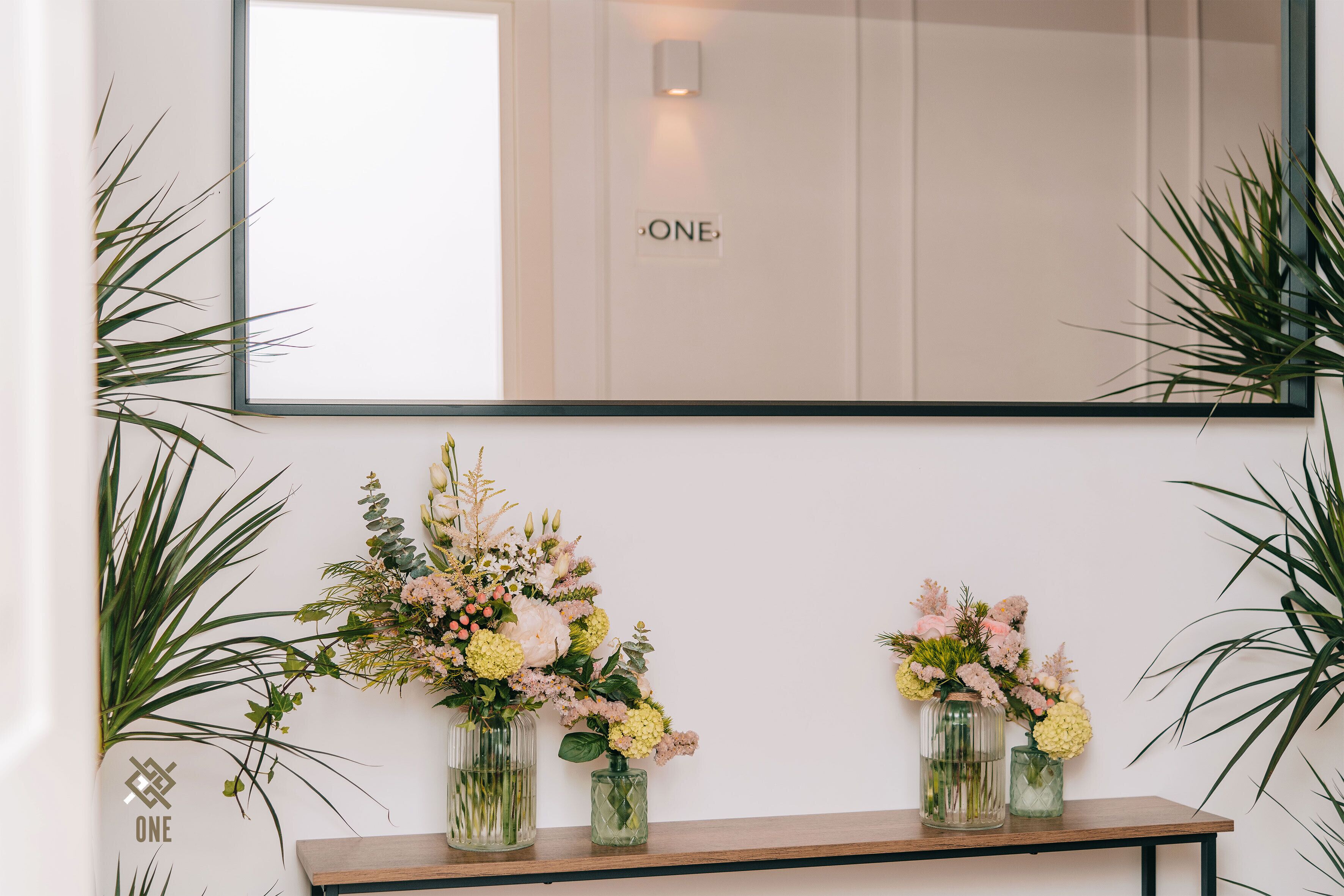 Ein Spiegel spiegelt ein Schild mit der Aufschrift 'ONE' an einer weißen Wand wider, mit zwei Glasvasen mit Blumen auf einem Holzregal.