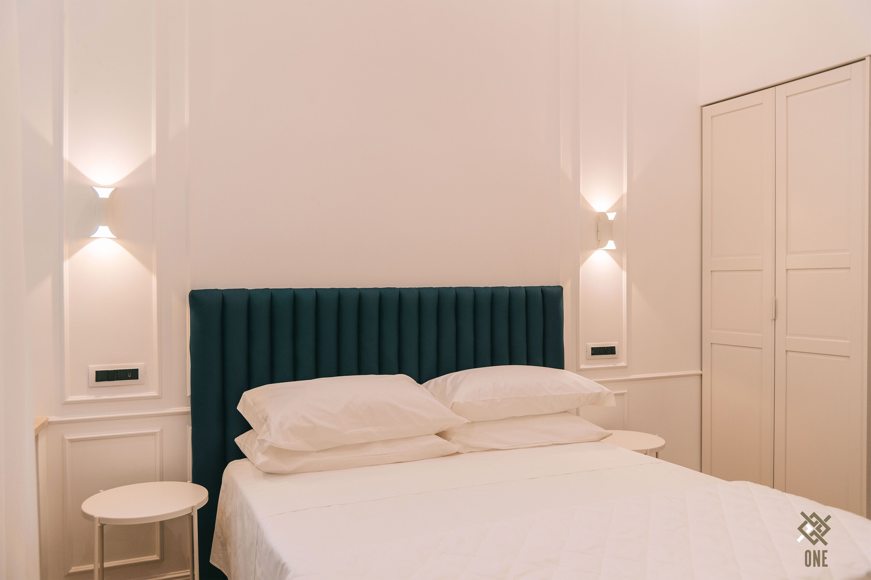A neatly made bed with white linens and pillows, a teal upholstered headboard, and wall-mounted sconces on either side.