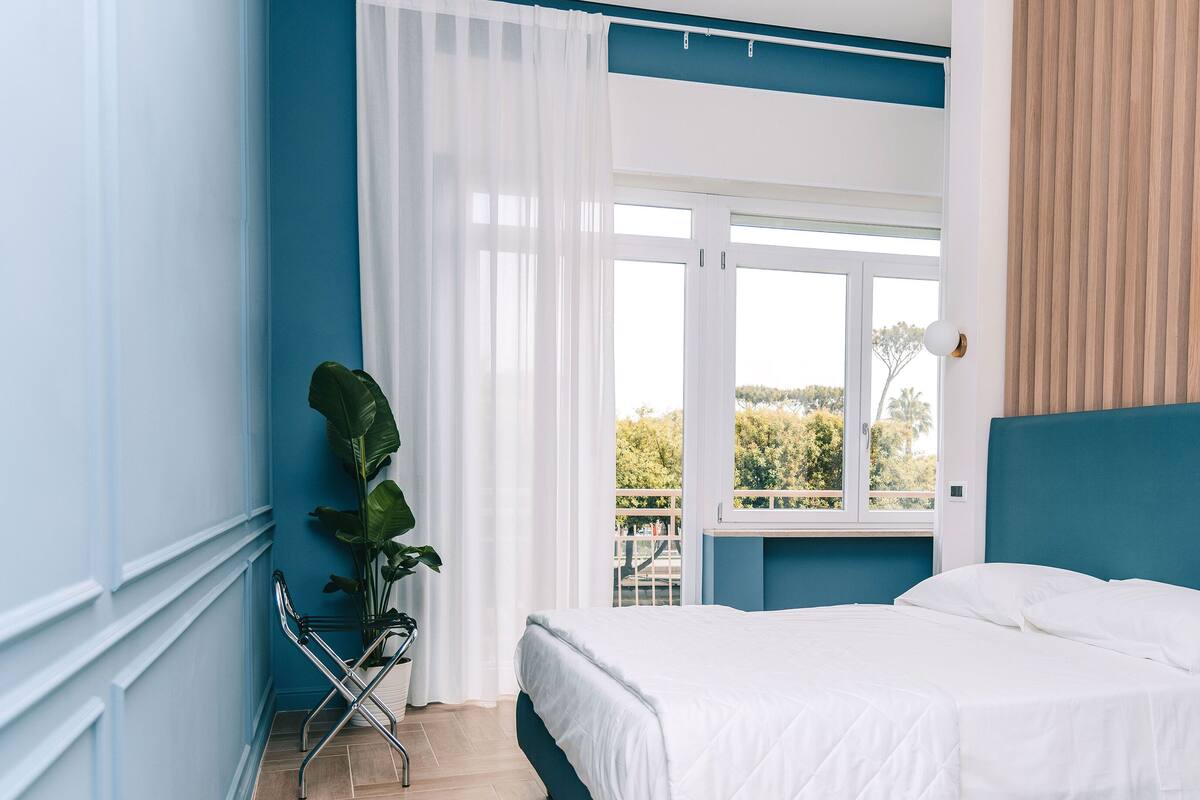 A bedroom with a bed, a window, a plant, and a view of the outdoors.