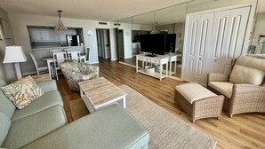 A modern living room with a sofa, ottoman, and wicker chairs, a dining area with chairs and a table, a kitchen in the background, and a TV on a stand. - New for June 2023 All New!! Taking Reservations Now w/Great Rates! (Myrtle Beach)