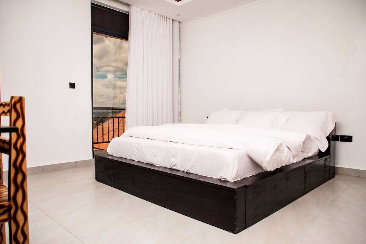 A modern bedroom with a large bed, a view of the sky through a window, and a balcony.