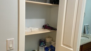 En delvis öppen garderob med hyllor som innehåller olika föremål, bland annat en dammsugare, en förpackning toalettpapper och städmaterial.