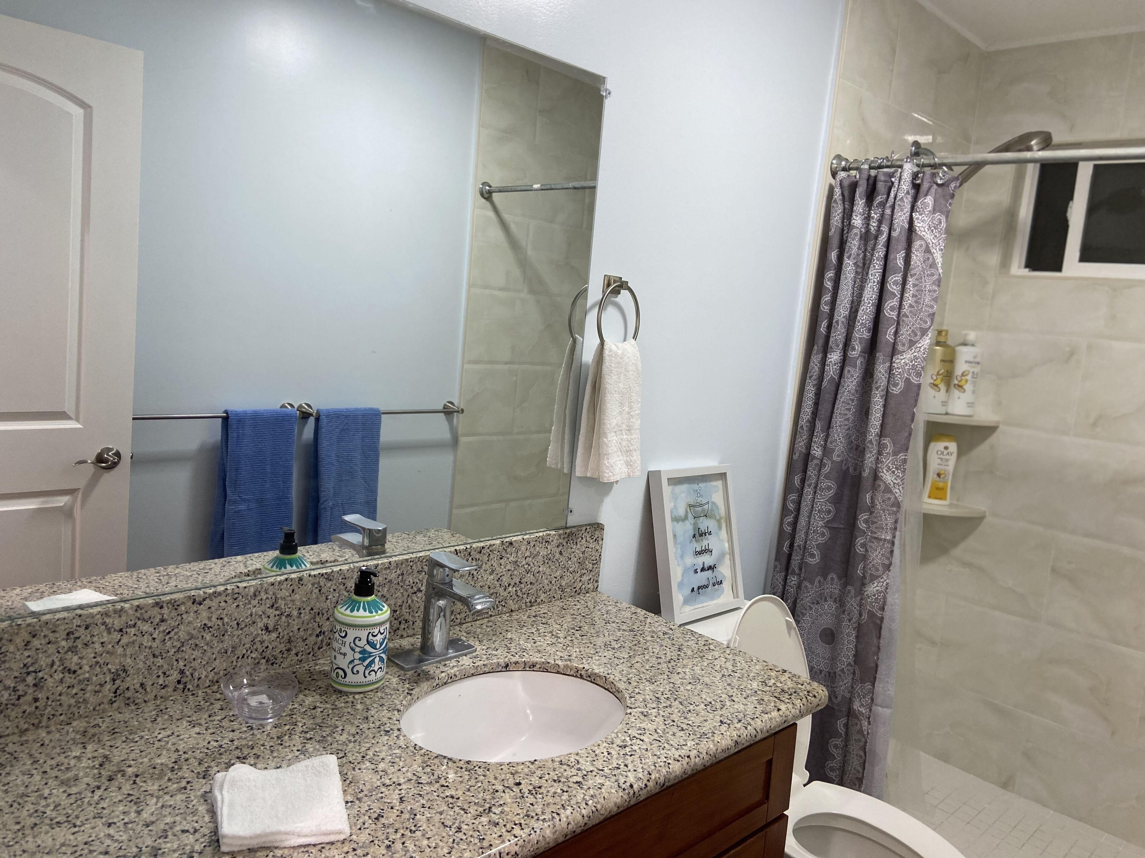 A bathroom with a granite countertop, a sink, a mirror, a towel rack, a shower curtain, and toiletries on a shelf.