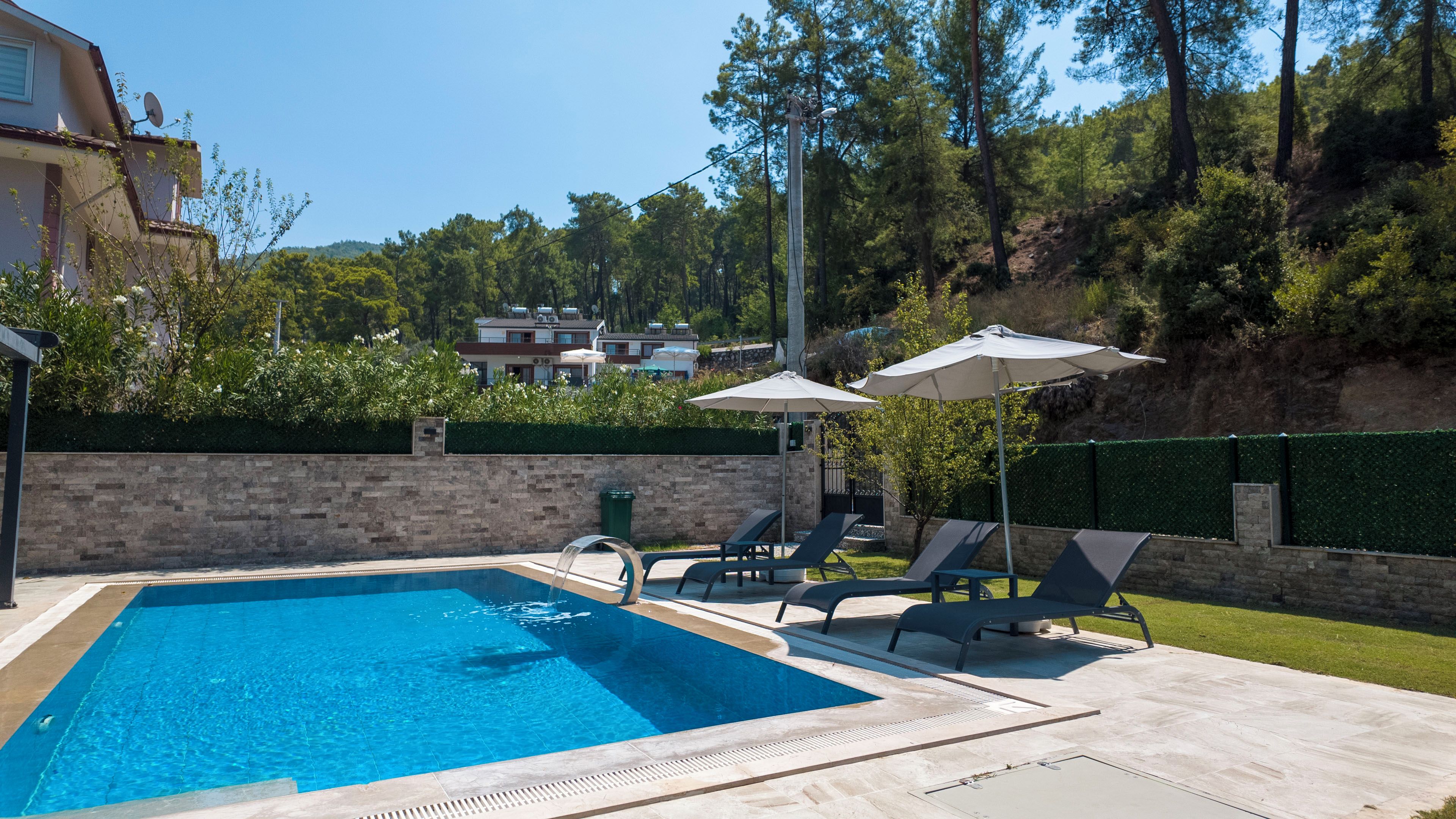 A swimming pool with lounge chairs and umbrellas, surrounded by a stone wall and greenery.