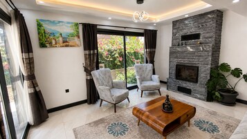 Una sala de estar con chimenea, dos sillones, una mesa de centro y un amplio ventanal con cortinas.