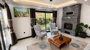 A living room with a fireplace, two armchairs, a coffee table, and a large window with curtains. - Villa Kayahan Göcek (Fethiye)