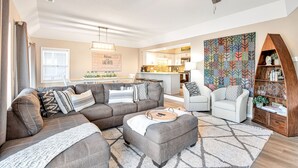Un salon moderne avec un canapé d’angle, un pouf et une table basse. On y trouve également un coin repas avec une table et des chaises, et une cuisine en arrière-plan.