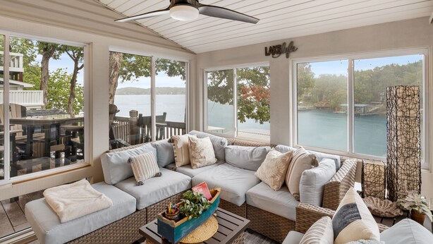 Une véranda avec une grande fenêtre donnant sur un lac, un ventilateur de plafond et un coin salon confortable avec des coussins et une table basse en osier.