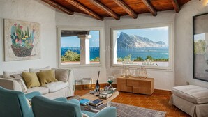 Un salón acogedor con techo de madera, un sofá blanco, sillones color turquesa y vistas al mar a través de grandes ventanales.