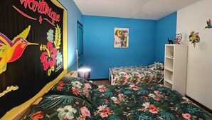 Quarto com parede azul, cama com colcha florida, mesa de cabeceira, prateleira na parede e pôster com um pássaro e flores.