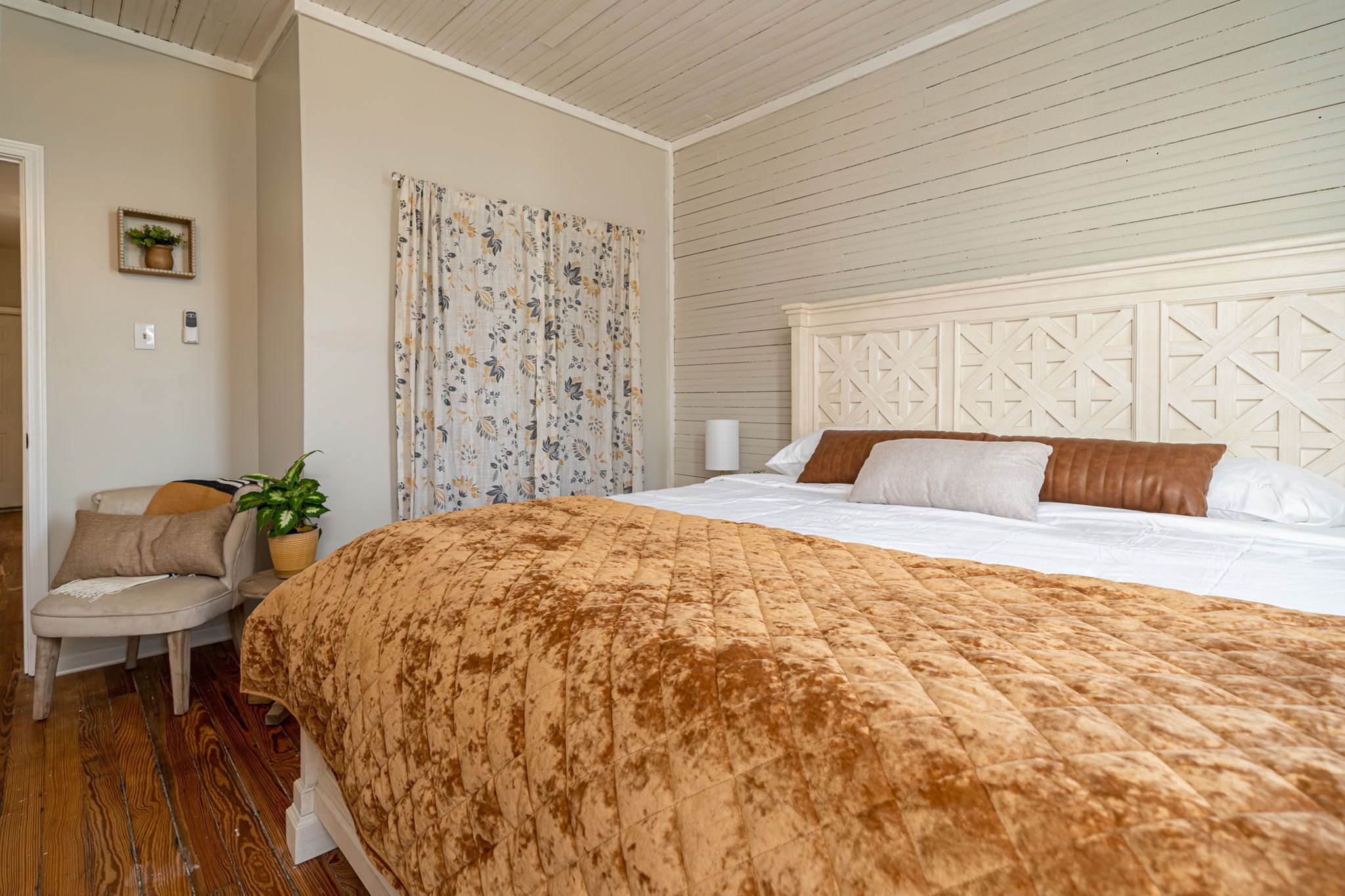 A bedroom with a large bed, a chair, a potted plant, and a wall-mounted light fixture.