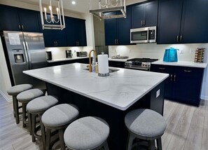 A modern kitchen with dark cabinets, a large island with stools, stainless appliances, and a chandelier.