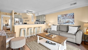 Una sala de estar moderna con un sofá gris, una mesa de centro de madera y una cocina con taburetes de bar.
