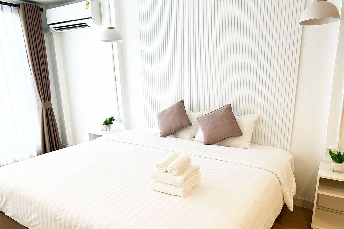 Una habitación de hotel con una cama grande, ropa de cama blanca, dos almohadas, una mesita de noche con una planta y un equipo de aire acondicionado.