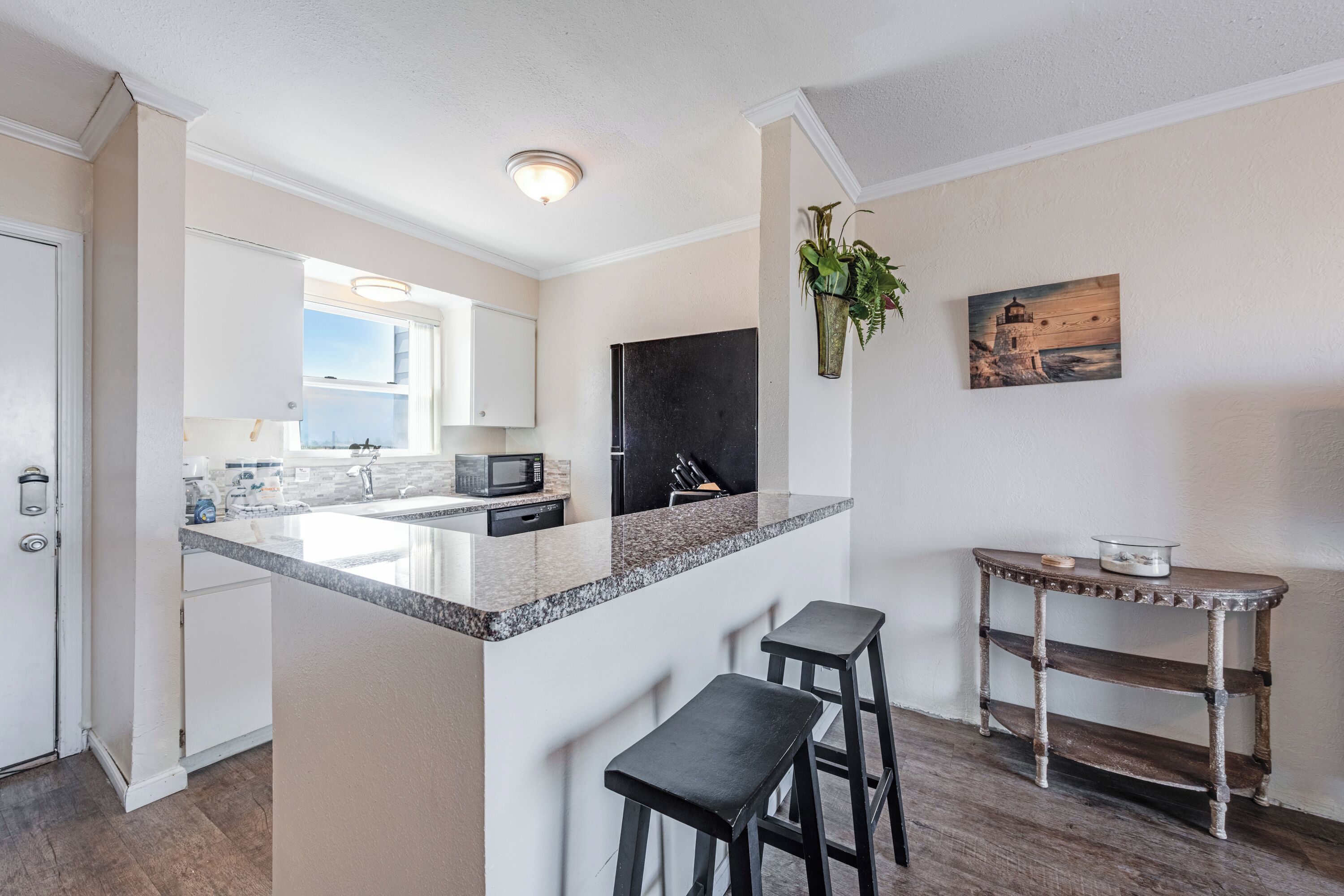 A modern kitchen with a granite countertop, white cabinets, and a black refrigerator. There are two bar stools and a small table with a plant.