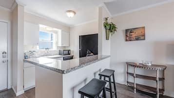 A modern kitchen with a granite countertop, white cabinets, and a black refrigerator. There are two bar stools and a small table with a plant.