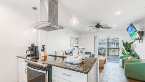 A modern kitchen with a central island, stainless steel appliances, and a dining area with a green sofa and a flat-screen TV. - With the best amenities in Port Aransas! Large swimming pool, private lighted fi (Port Aransas)