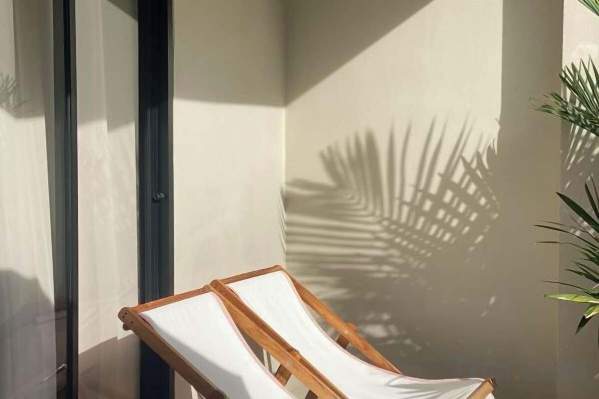 A sunlit room with a window, a wooden chair, and a shadow of a palm tree on the wall.